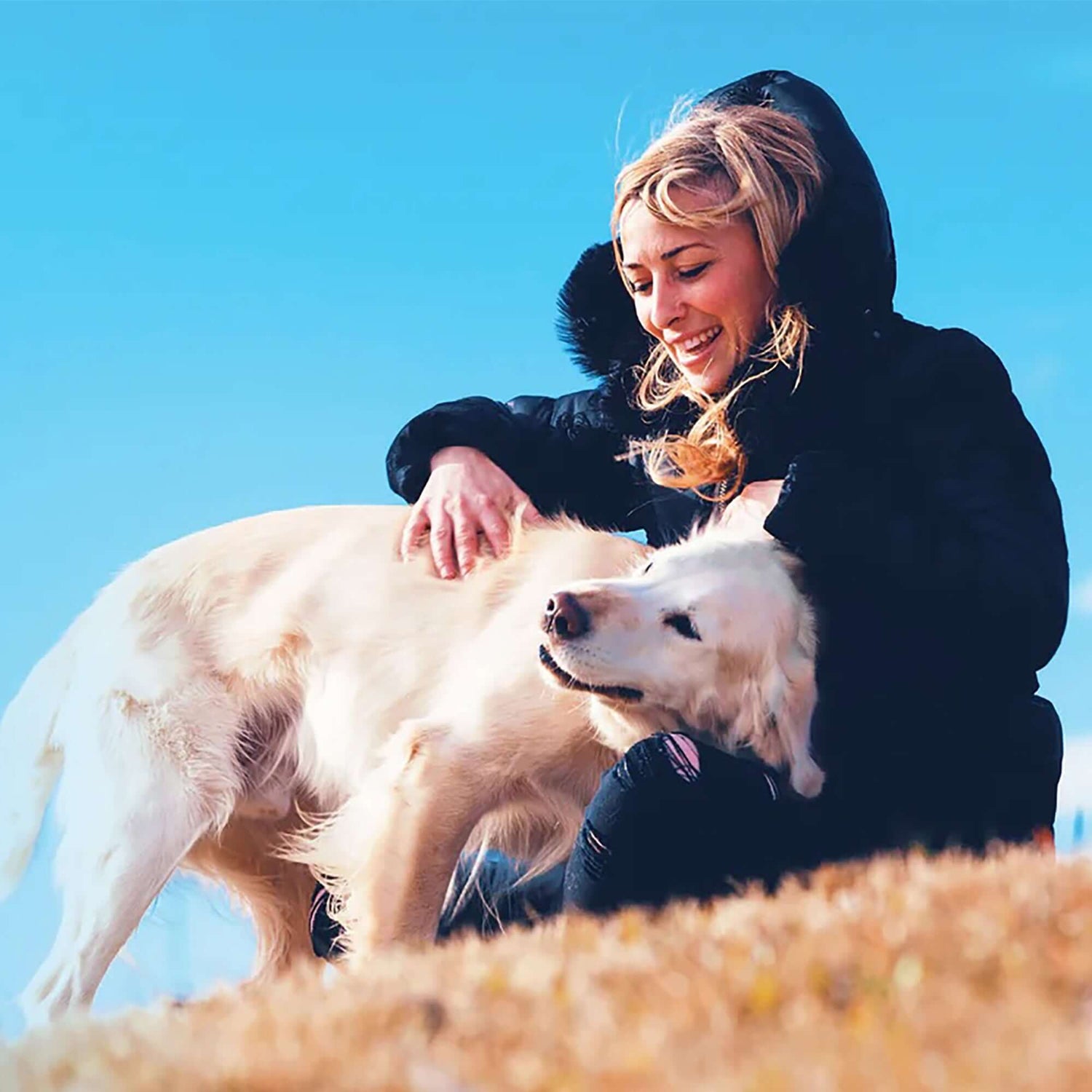 Woman with blonde hair petting a golden retriever on a sunny hillside.