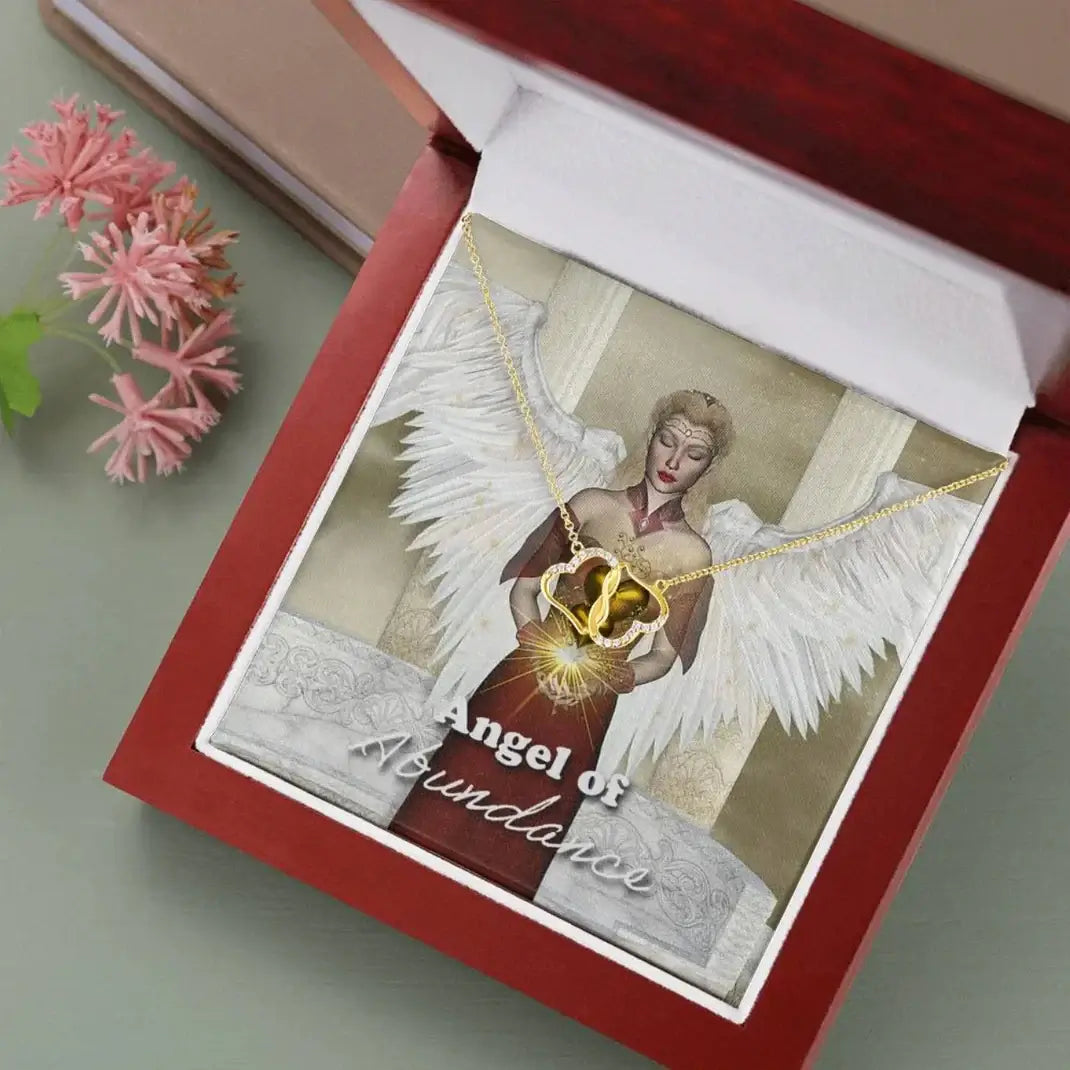 Angel of Abundance necklace with angel wings design displayed in an elegant gift box with pink flowers nearby.