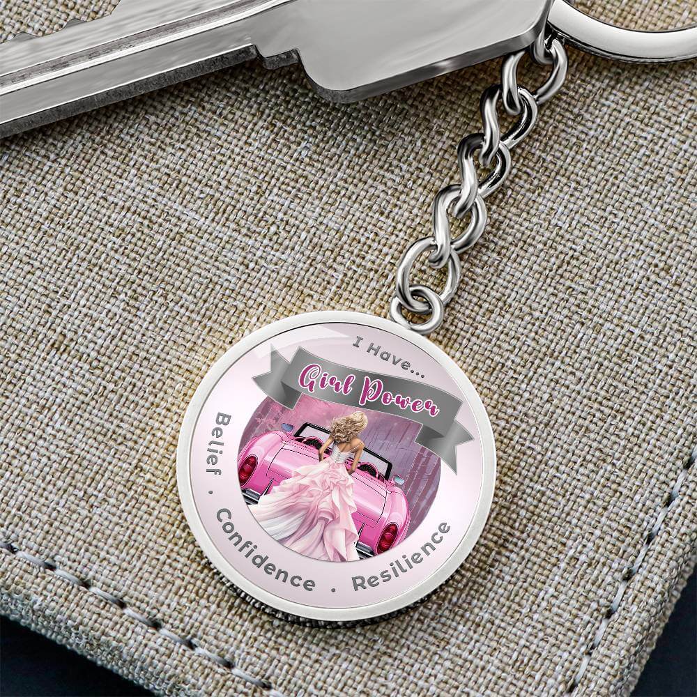 Keychain with a "Girl Power" design featuring a pink car and inspirational words: Belief, Confidence, Resilience, on a fabric surface.
